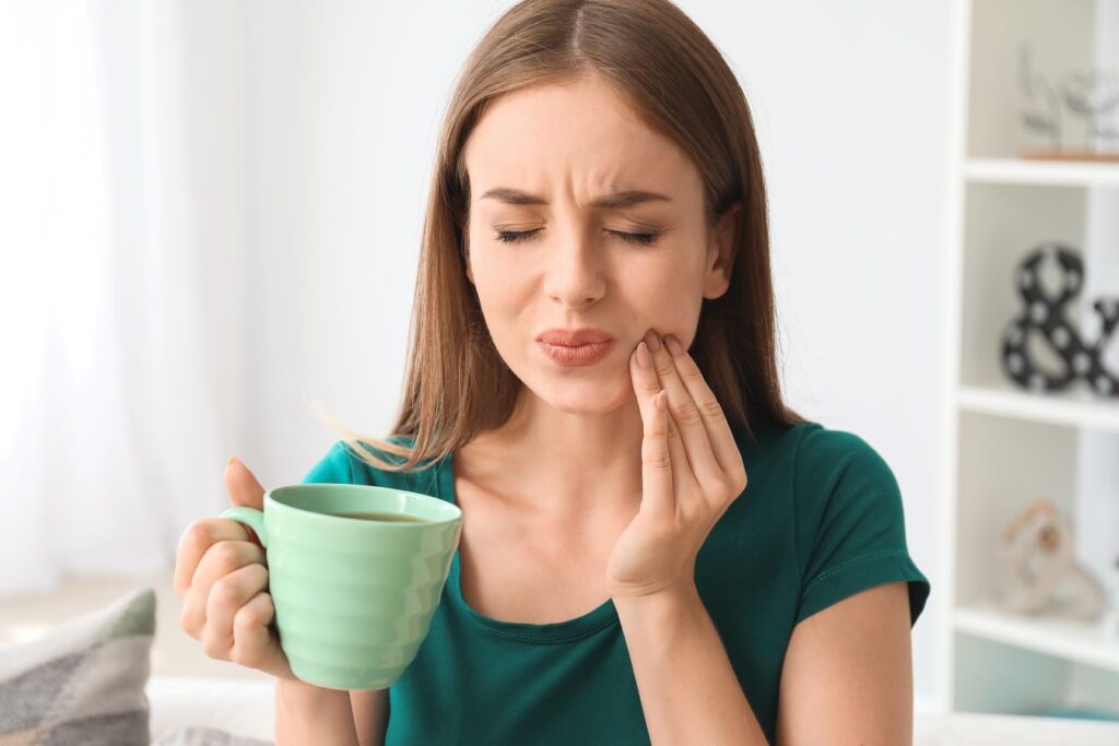 A woman holding a cup of coffee and touching her face in pain from tooth sensitivity.